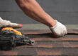 closeup of roofer placing new shingles on a roof with nail gun laying nearby.