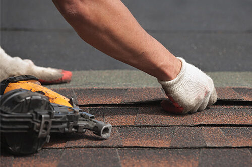 closeup of roofer placing new shingles on a roof with nail gun laying nearby.
