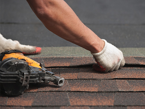 closeup of roofer placing new shingles on a roof with nail gun laying nearby.